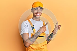 Young handsome african american man wearing handyman uniform over yellow background smiling and looking at the camera pointing