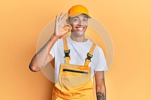 Young handsome african american man wearing handyman uniform over yellow background smiling positive doing ok sign with hand and