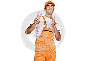 Young handsome african american man wearing handyman uniform smiling looking to the camera showing fingers doing victory sign