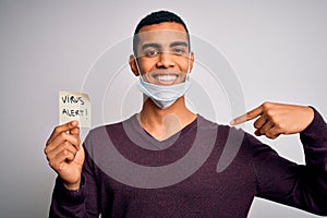 Young handsome african american man wearing medical mask holding virus alert reminder with surprise face pointing finger to