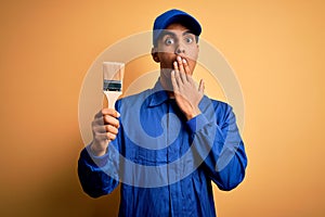 Young handsome african american painter man wearing uniform using painting brush cover mouth with hand shocked with shame for