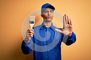 Young handsome african american painter man wearing uniform using painting brush with open hand doing stop sign with serious and