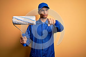 Young handsome african american painter man wearing uniform using painting roller pointing with finger to the camera and to you,