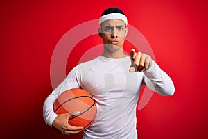 Young handsome african american sportsman holding basketball ball over red background pointing with finger to the camera and to