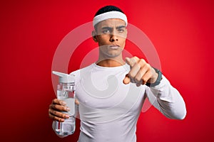 Young handsome african american sportsman wearing sportswear drinking bottle of water pointing with finger to the camera and to