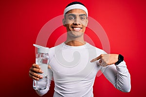 Young handsome african american sportsman wearing sportswear drinking bottle of water with surprise face pointing finger to