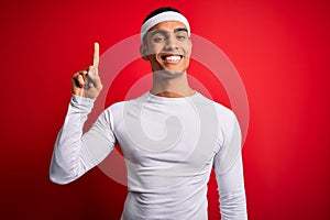 Young handsome african american sportsman wearing sportswear over red background showing and pointing up with finger number one