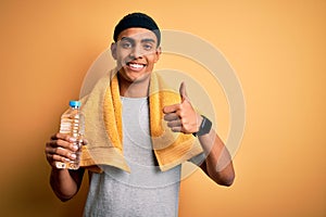 Young handsome african american sportsman wearing towel drinking bottle of water happy with big smile doing ok sign, thumb up with