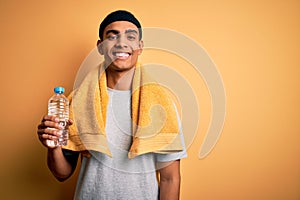 Young handsome african american sportsman wearing towel drinking bottle of water with a happy face standing and smiling with a