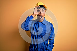 Young handsome african american worker man wearing blue uniform and security helmet doing ok gesture shocked with surprised face,
