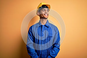 Young handsome african american worker man wearing blue uniform and security helmet looking away to side with smile on face,