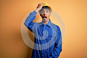 Young handsome african american worker man wearing blue uniform and security helmet surprised with hand on head for mistake,