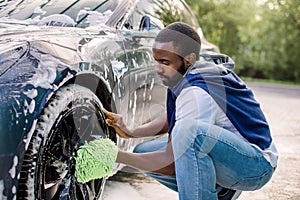 Young handsome African guy holding green sponge, washing car wheel with foam. Cleaning of modern rims of luxury blue