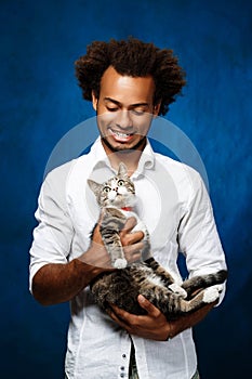 Young handsome african man holding cat over blue background.