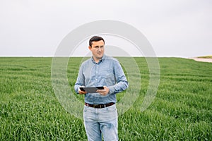 Young handsome agronomist, agriculture engineer standing in green wheat field with tablet in hands in early summer. Agribusiness