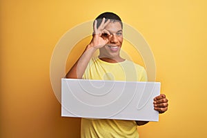 Young handsome arab man holding banner standing over isolated yellow background with happy face smiling doing ok sign with hand on