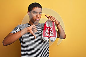 Young handsome arab man holding casual sneakers standing over isolated yellow background with angry face, negative sign showing