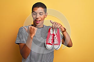 Young handsome arab man holding casual sneakers standing over isolated yellow background pointing and showing with thumb up to the