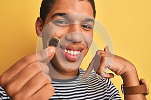 Young handsome arab man holding euro coin standing over isolated yellow background with surprise face pointing finger to himself