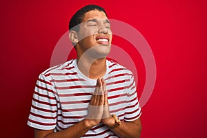 Young handsome arab man wearing striped t-shirt over isolated red background begging and praying with hands together with hope