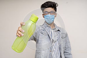 Young handsome asian boy in denims, wearing a protective mask, holding a water bottle. Confident boy showing to drink more water