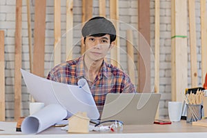 Young handsome asian male architect with black hair wearing plaid shirt sit and work behind working desk with some drawing
