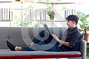 Young handsome Asian man in black shirt typing on keyboard laptop computer while lying down comfortably on sofa couch in his