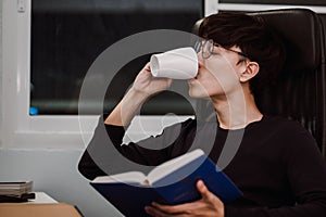 Young handsome asian man drinking tea to relax from heavy reading book at work desk late at night, Knowledge and learning concept.