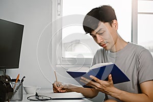 Young handsome asian man reading book and writing in note at work desk in free time from working at home, Knowledge and learning