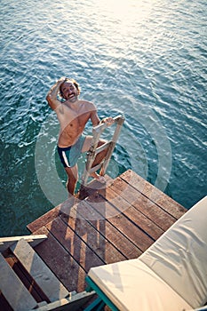 Young handsome athletic man in swimsuit shorts comming out of water, up on ladder to wooden jetty. Man on summer vacation by sea. photo