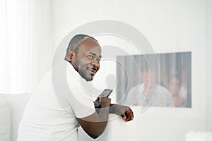 Young handsome and attractive black african american man sitting at home sofa couch watching television using tv remote controller