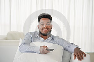Young handsome and attractive black african american man sitting at home sofa couch watching television using tv remote controller