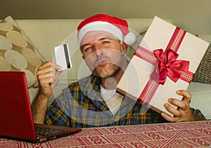 Young handsome and attractive happy man in Santa hat at home couch using laptop computer and credit card holding Christmas gift bo