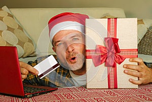 Young handsome and attractive happy man in Santa hat at home couch using laptop computer and credit card holding Christmas gift bo
