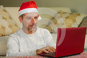 Young handsome and attractive happy man in Santa hat at home online shopping on internet with laptop computer using credit card bu