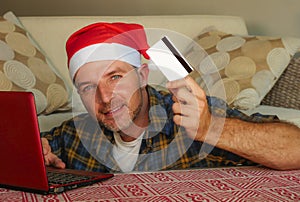 Young handsome and attractive happy man in Santa hat at home online shopping on internet with laptop computer using credit card bu