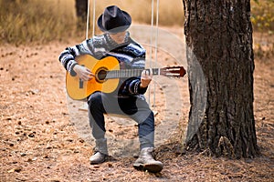 Young handsome attractive man in sweater playing guitar in forest sitting on swing. Casual man, life style