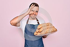 Young handsome baker man with blue eyes wearing apron holding tray with wholmeal bread with happy face smiling doing ok sign with