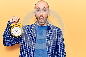 Young handsome bald man holding alarm clock scared and amazed with open mouth for surprise, disbelief face