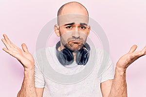 Young handsome bald man listening to music using headphones clueless and confused with open arms, no idea and doubtful face