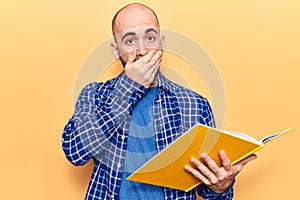 Young handsome bald man reading book covering mouth with hand, shocked and afraid for mistake
