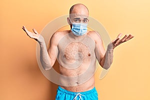 Young handsome bald man shirtless wearing swimwear and medical mask clueless and confused with open arms, no idea and doubtful