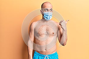 Young handsome bald man shirtless wearing swimwear and medical mask pointing thumb up to the side smiling happy with open mouth