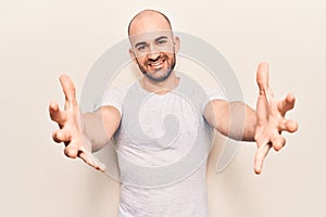 Young handsome bald man wearing casual t shirt looking at the camera smiling with open arms for hug