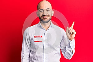 Young handsome bald man wearing hello my name is sticker smiling happy pointing with hand and finger to the side