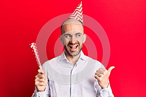 Young handsome bald man wearing party hat and trumpet pointing thumb up to the side smiling happy with open mouth