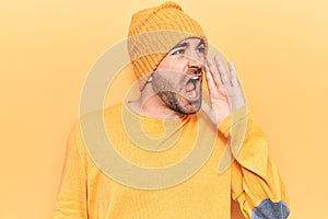 Young handsome bald man wearing sweater and wool cap shouting and screaming loud to side with hand on mouth