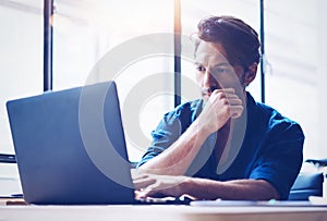 Young handsome banking finance analyst working at sunny office on laptop while sitting at wooden table.Businessman