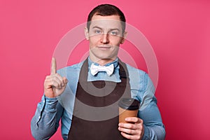 Young handsome barista man, stands enjoying cup of coffee isolated on pink background, points up with index figer. Attractive male