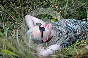 A young handsome bearded blonde guy wearing black sunglasses lying on the grass and looking up to sky.